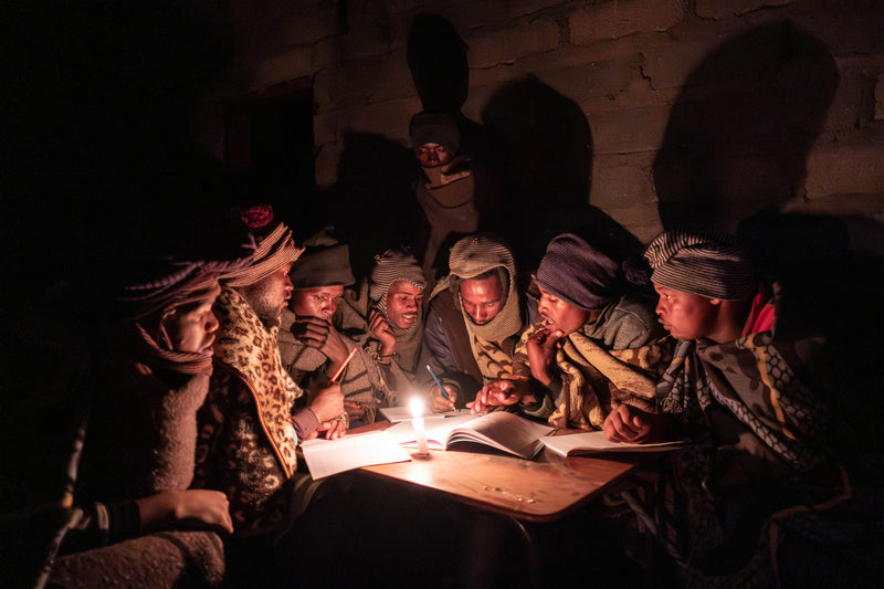 Lesotho herders gathered around a solar‑lit night‑school table reading basic alphabet cards